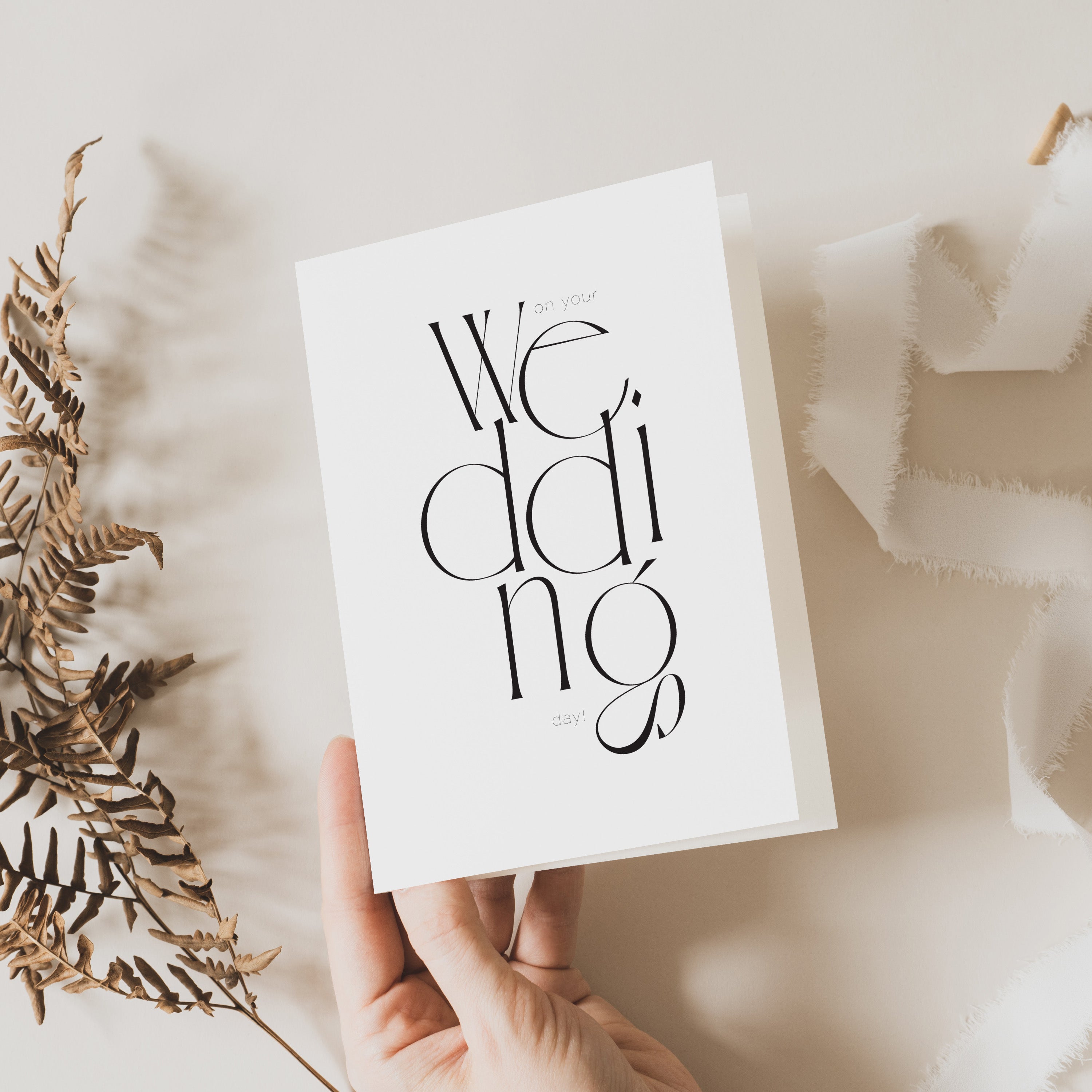 A hand holding a modern and abstract wedding day card with the word 'wedding' written in bold black letters, presented on a neutral background with some foliage decoration.