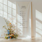 Cotton and linen wedding order of the day sign hanging on a stand with floral arrangement