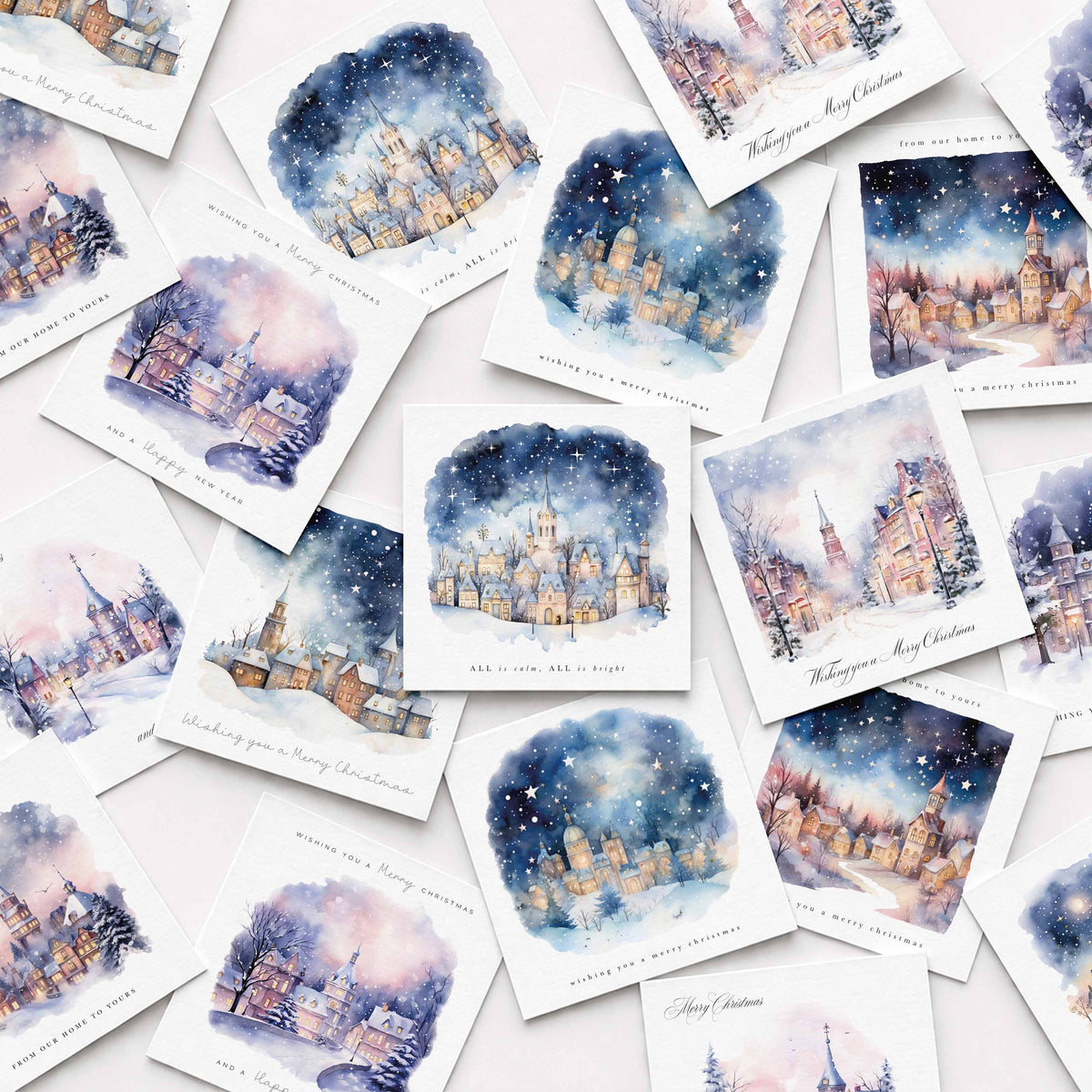 A flat-lay arrangement of multiple watercolor Christmas cards featuring snowy village scenes with warm glowing windows under a starry night sky, on a light pink background.