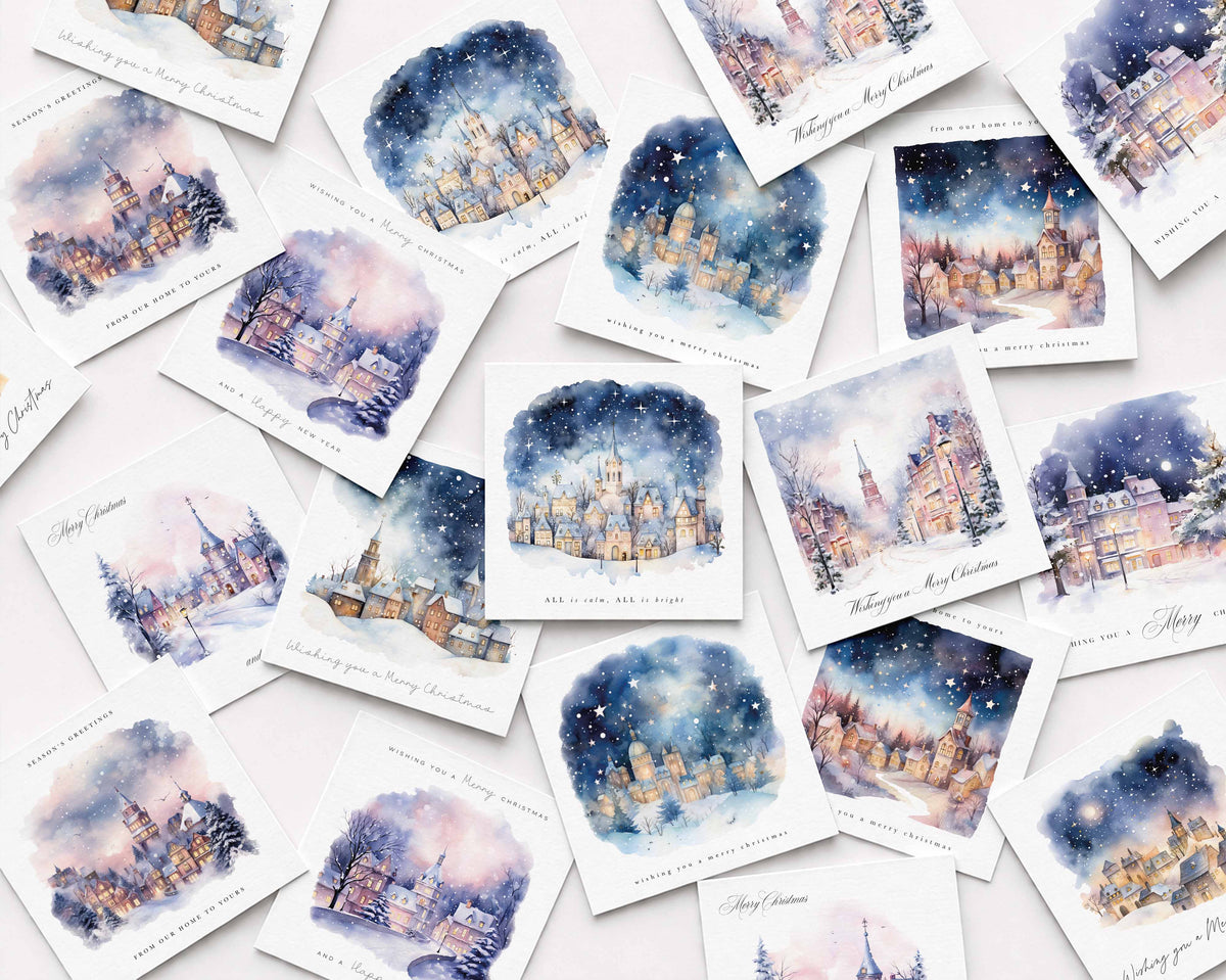 A flat-lay arrangement of multiple watercolor Christmas cards featuring snowy village scenes with warm glowing windows under a starry night sky, on a light pink background.