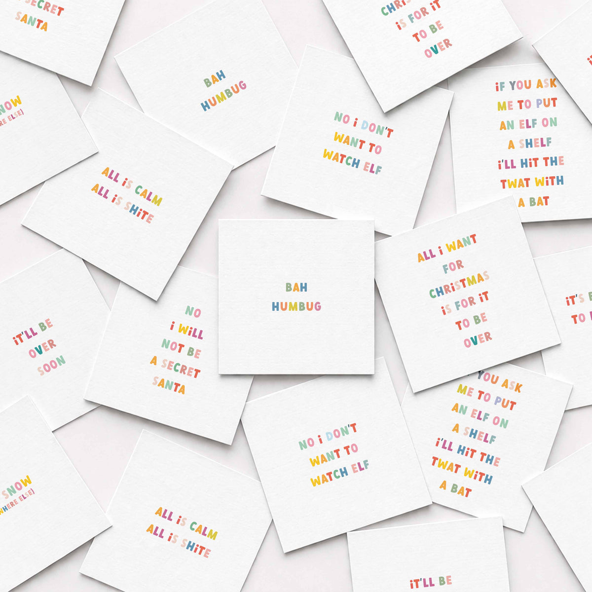 A scattered arrangement of small white square anti-Christmas cards printed with colorful playful text on a white background.