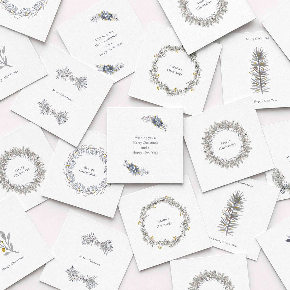 A scattered arrangement of white Christmas cards featuring delicate botanical illustrations in muted gray, blue, and yellow, including wreaths, sprigs, and small text such as Merry Christmas and Happy New Year.