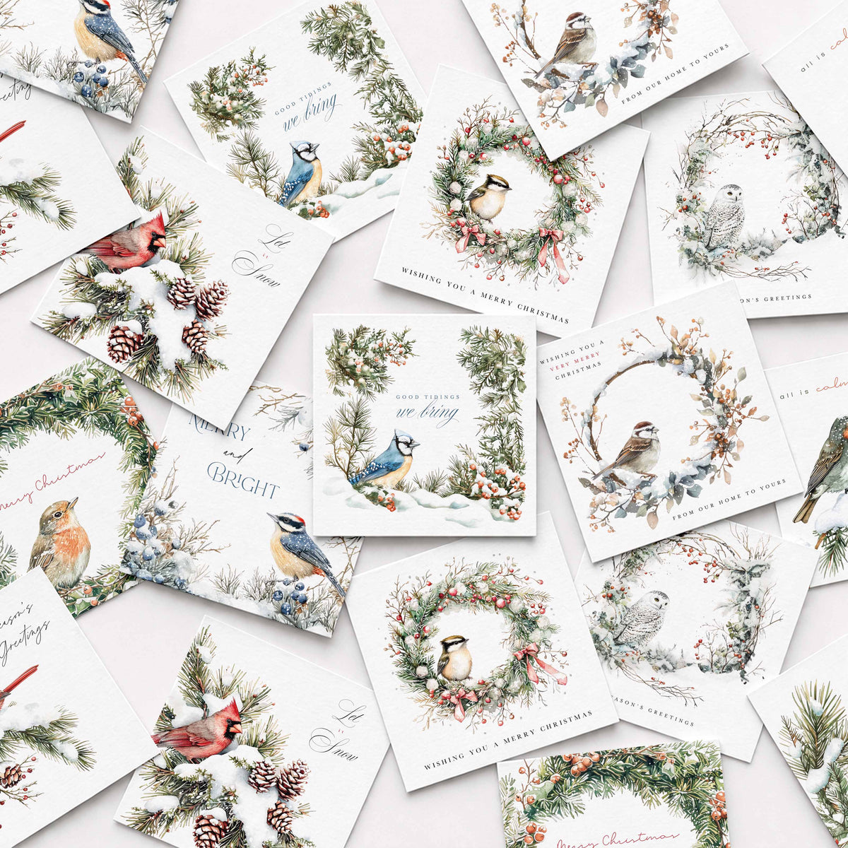 A flat-lay arrangement of multiple white Christmas greeting cards featuring watercolor illustrations of birds, wreaths, pine branches, and holiday text, photographed on a pale pink background.