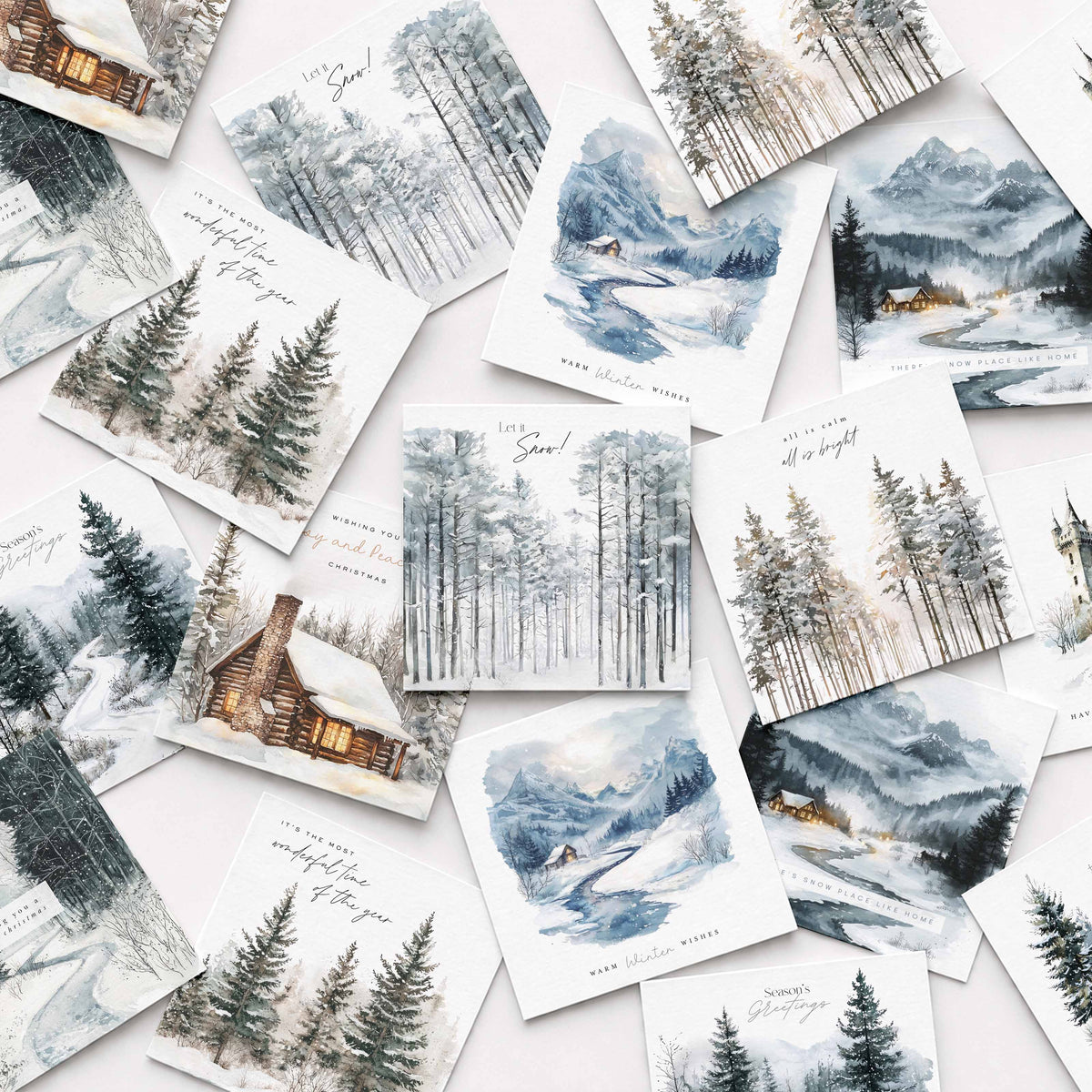 A flat-lay arrangement of multiple watercolor winter landscape Christmas cards scattered on a light background. The prints depict snowy forests, mountains, cabins, and rivers in soft blue, gray, white, and brown tones, with some cards featuring handwritten text and small holiday greetings.