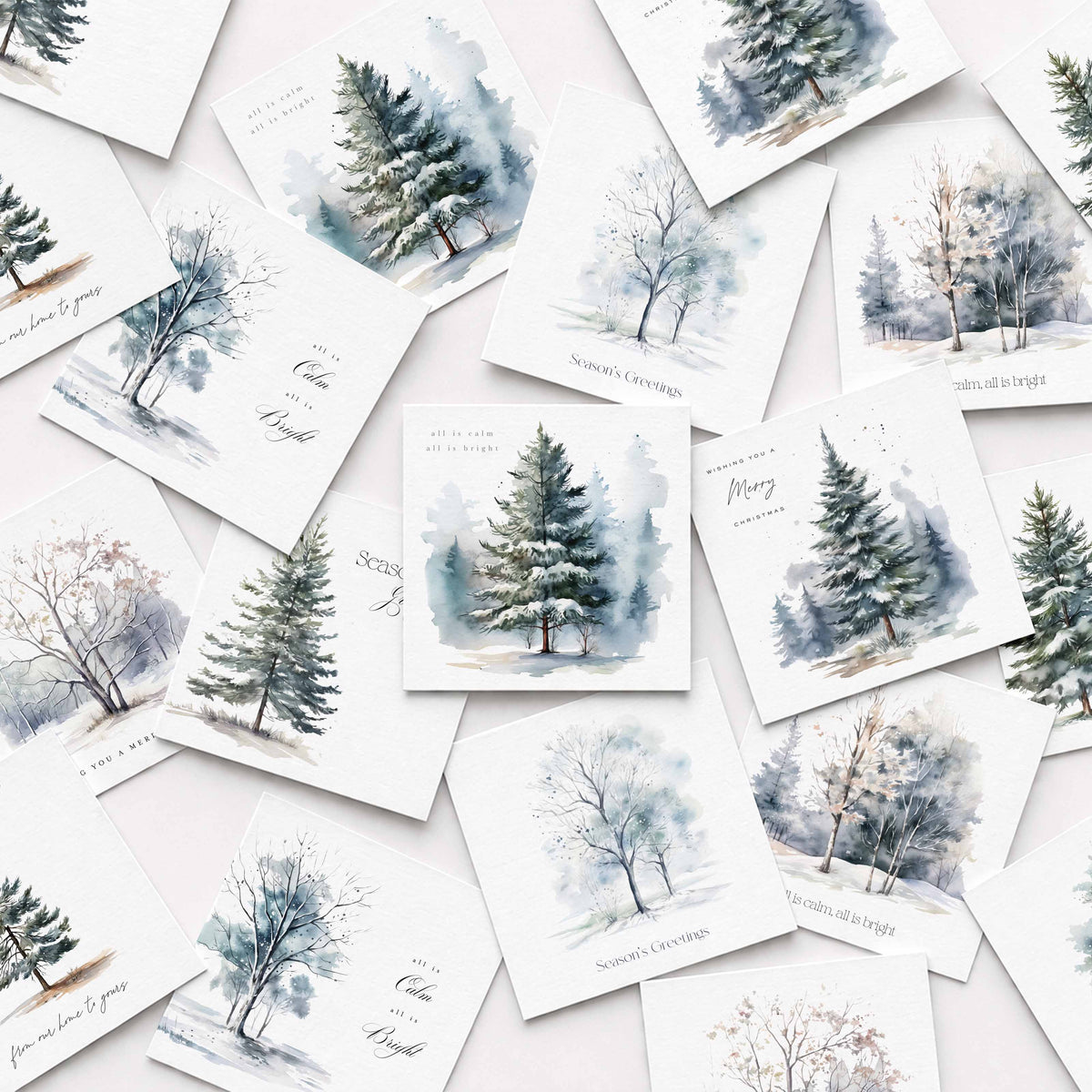 A flat-lay arrangement of multiple Christmas cards featuring watercolor winter trees and snowy forest scenes in soft blue, gray, and green tones on white backgrounds.