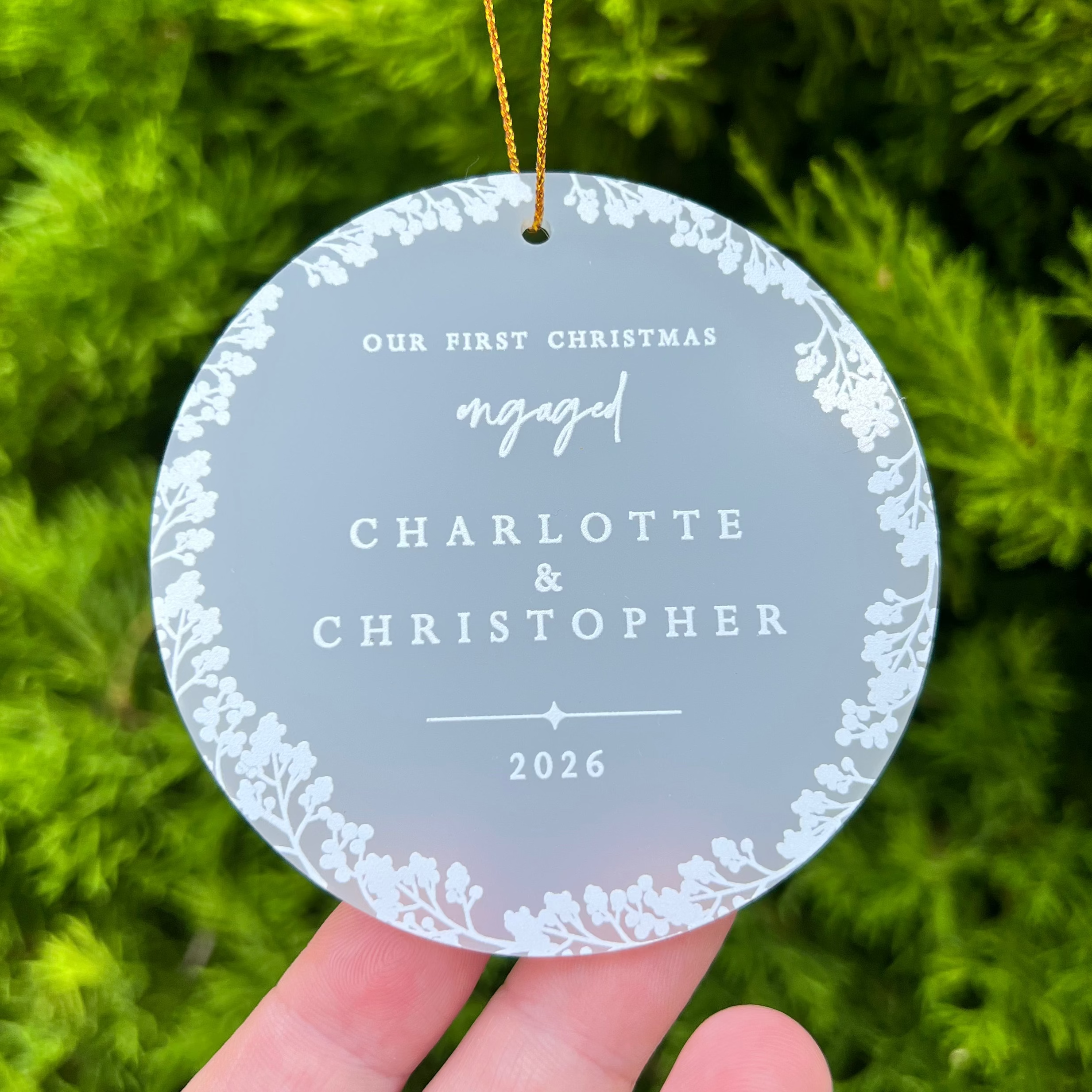 A round personalized Christmas ornament held in a hand against a background of green pine needles. The ornament is frosted acrylic with a white floral border and white text reading 'Our First Christmas engaged Charlotte & Christopher 2026' with a gold hanging string.
