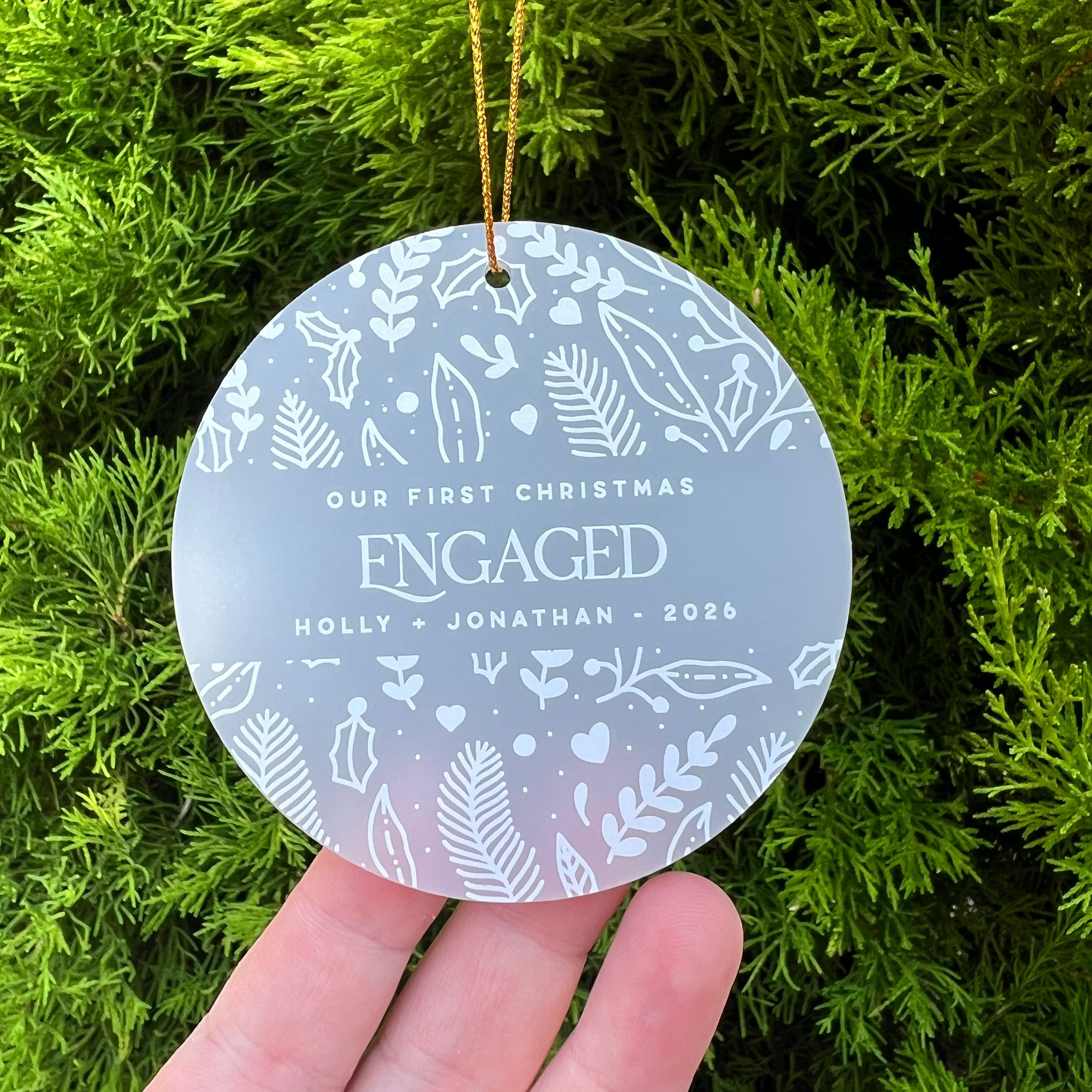 A round personalized Christmas ornament held in a hand against a background of green pine branches. The ornament is frosted light gray with white leaf and heart patterns and reads 'OUR FIRST CHRISTMAS ENGAGED HOLLY + JONATHAN - 2026'.