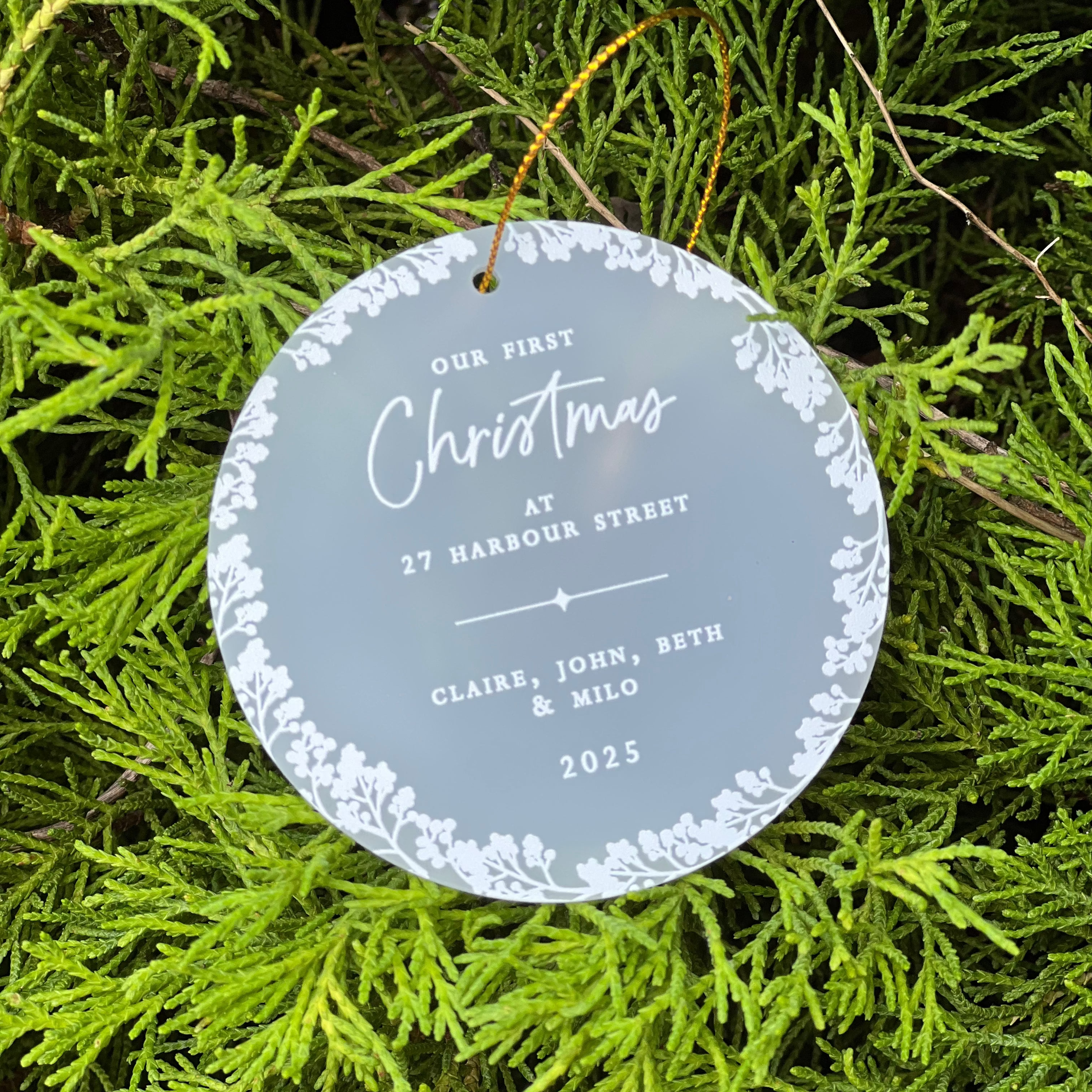 A round frosted glass Christmas ornament with white floral border and personalized text reading 'Our First Christmas at 27 Harbour Street' and names, hanging from a gold string amidst bright green pine branches.