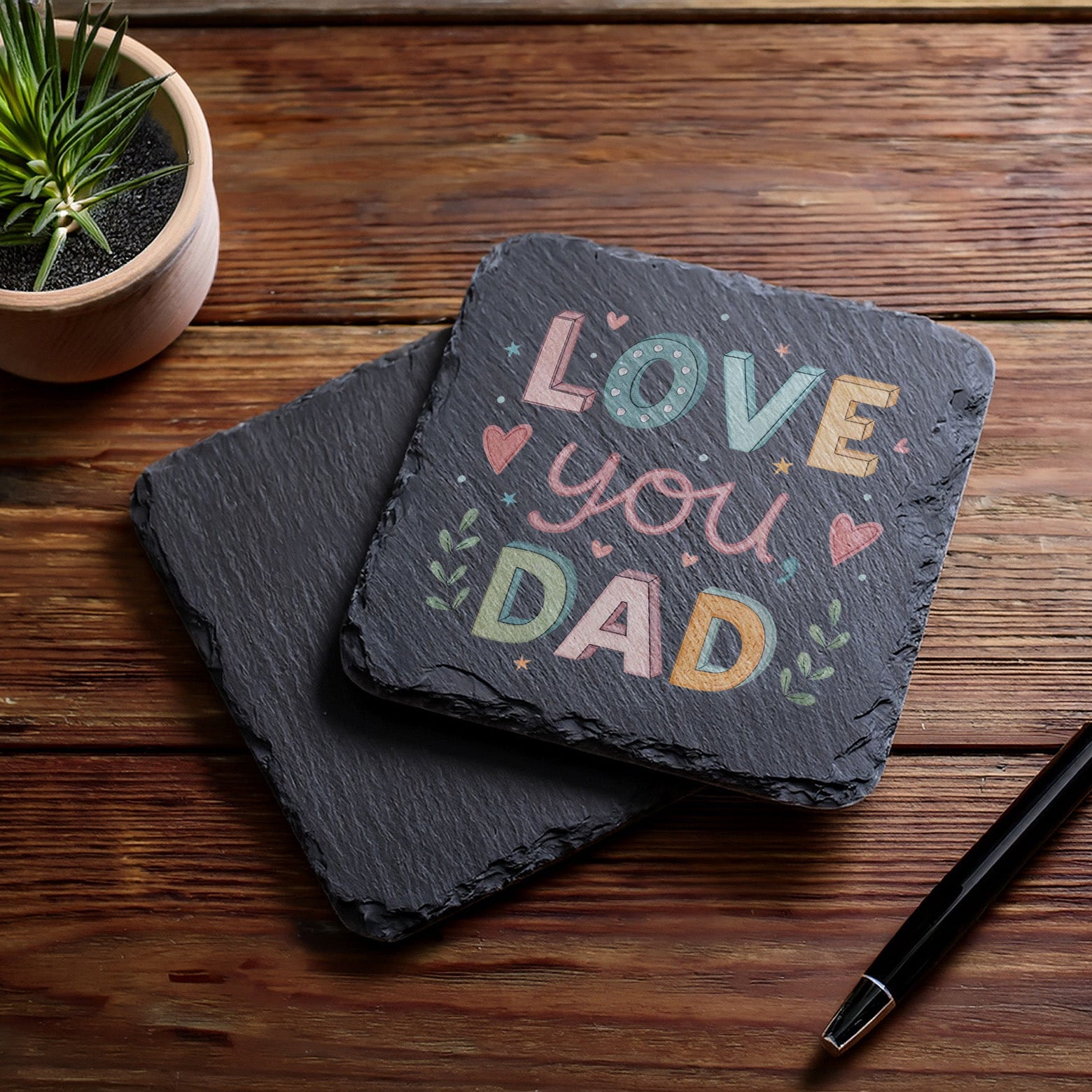 Slate coaster with 'Love you Dad' text on a wooden surface with a plant and pen.