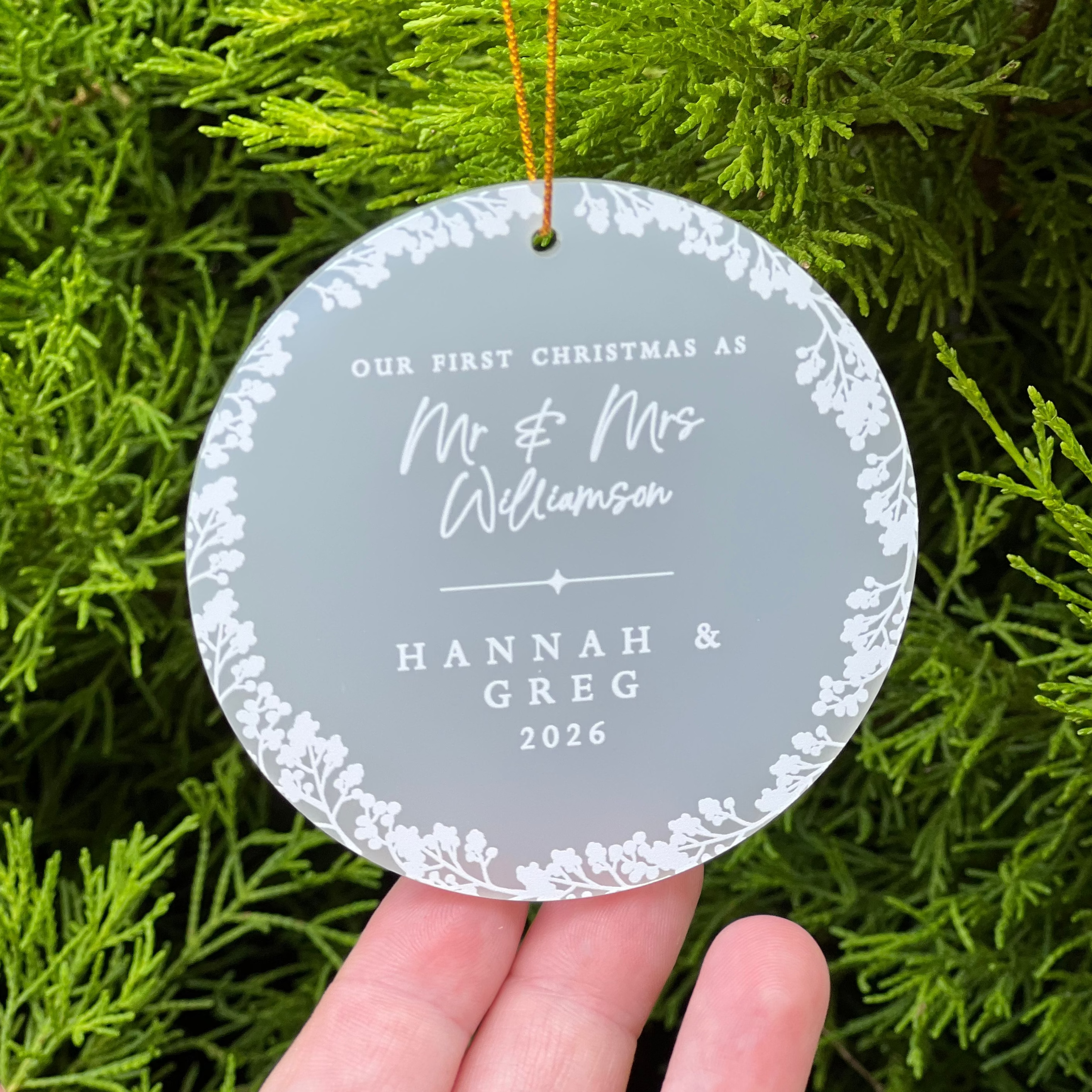 A personalized round Christmas ornament held in a hand against a background of green pine branches. The ornament is frosted acrylic with a white floral border and custom text reading 'OUR FIRST CHRISTMAS AS Mr & MRS WILLIAMSON"