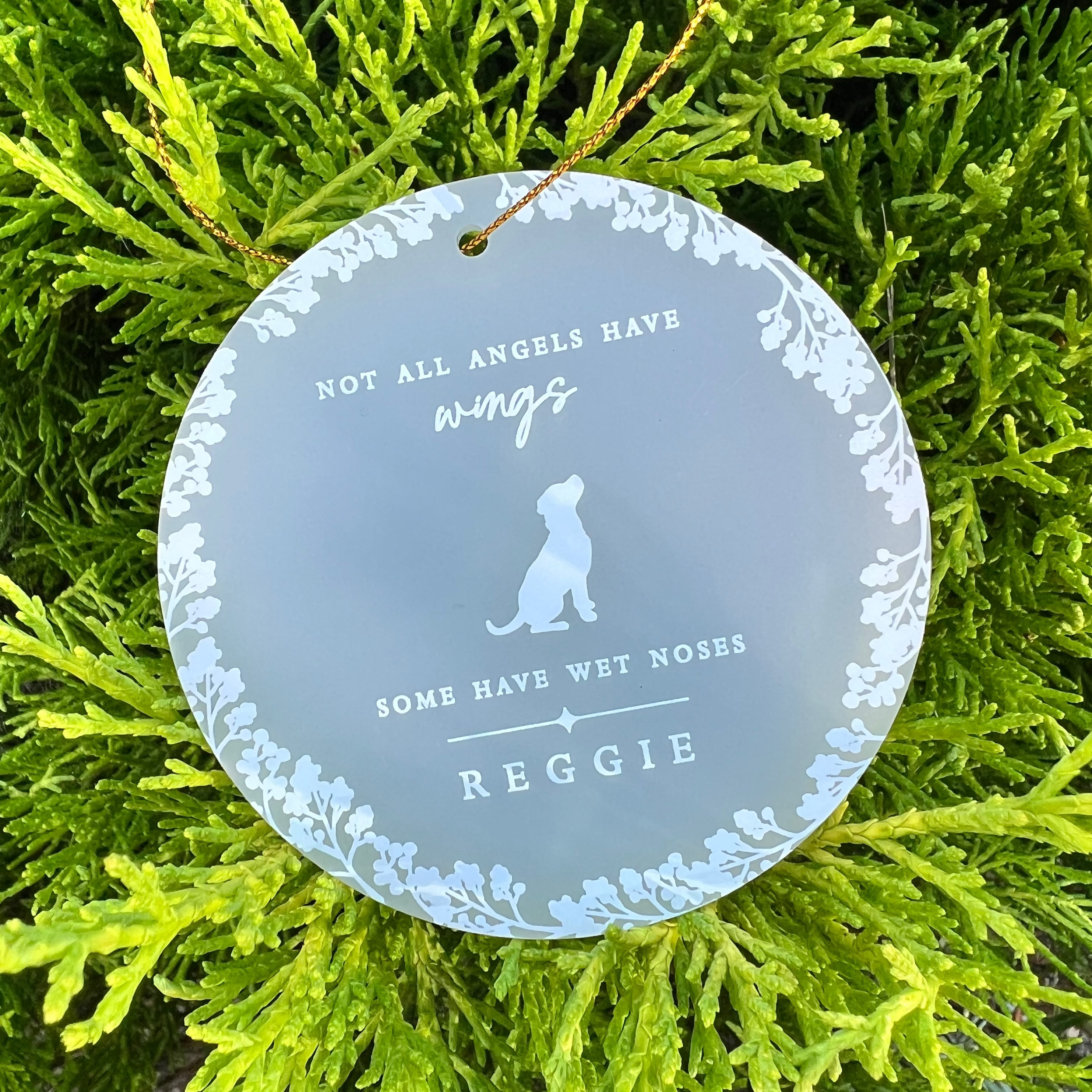 A round frosted acrylic pet memorial hanging ornament with white floral border and text reading 'NOT ALL ANGELS HAVE wings SOME HAVE WET NOSES REGGIE' and a dog silhouette, hanging from a tree branch.