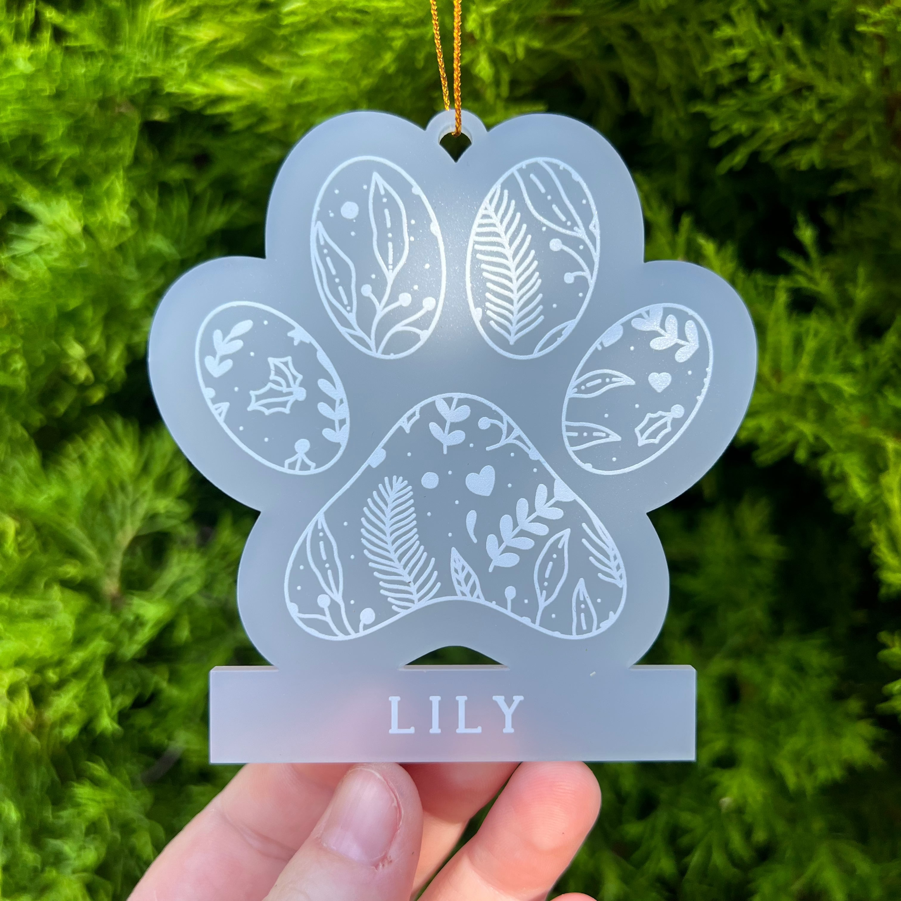 A frosted acrylic paw-shaped hanging ornament with white floral and heart patterns, personalized with the name 'LILY' on a base, held by a hand against a green pine background.