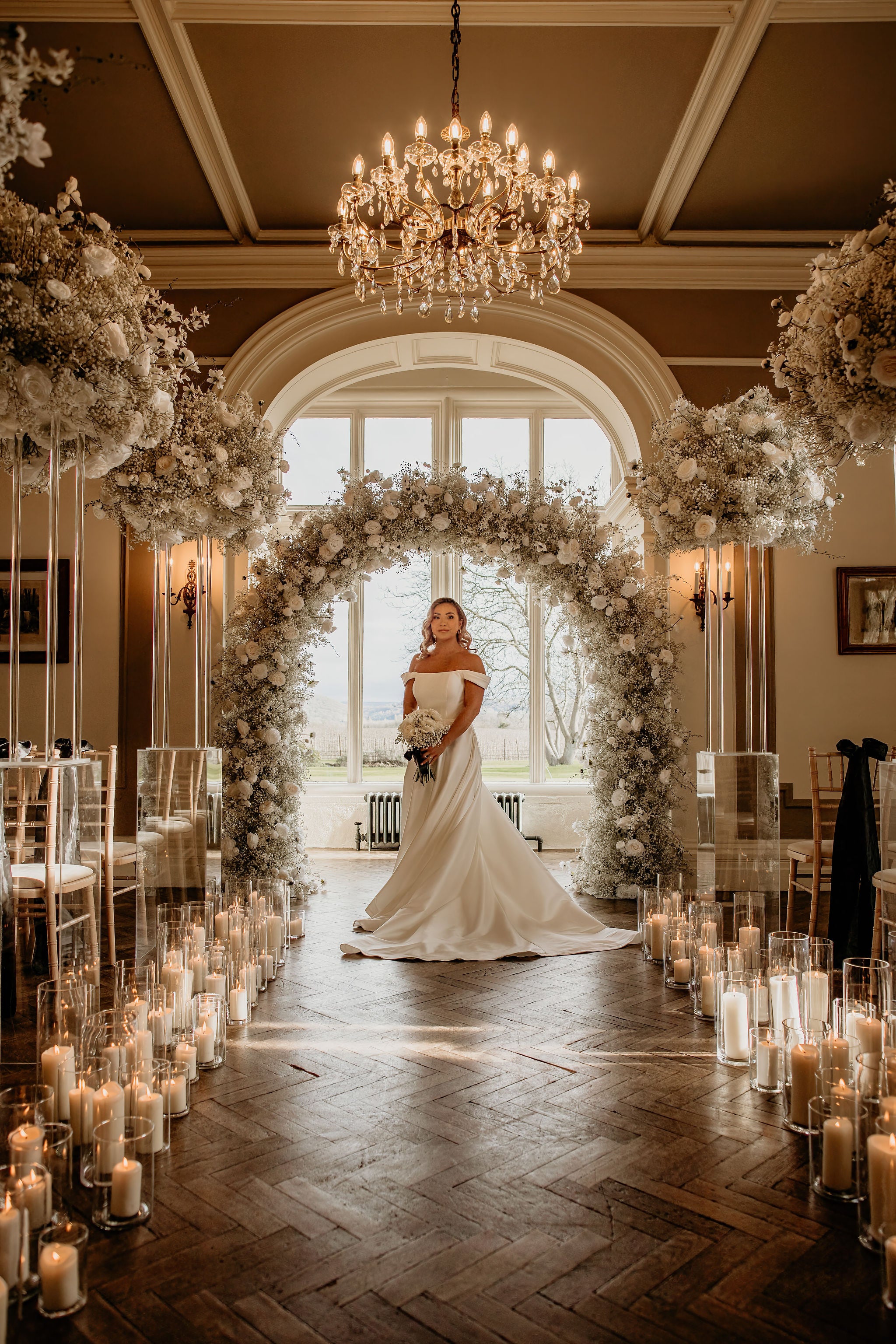 Black and white wedding arch, with bridge holding a white bouquet in a large statley home in front of a large indown, with candles down the isle