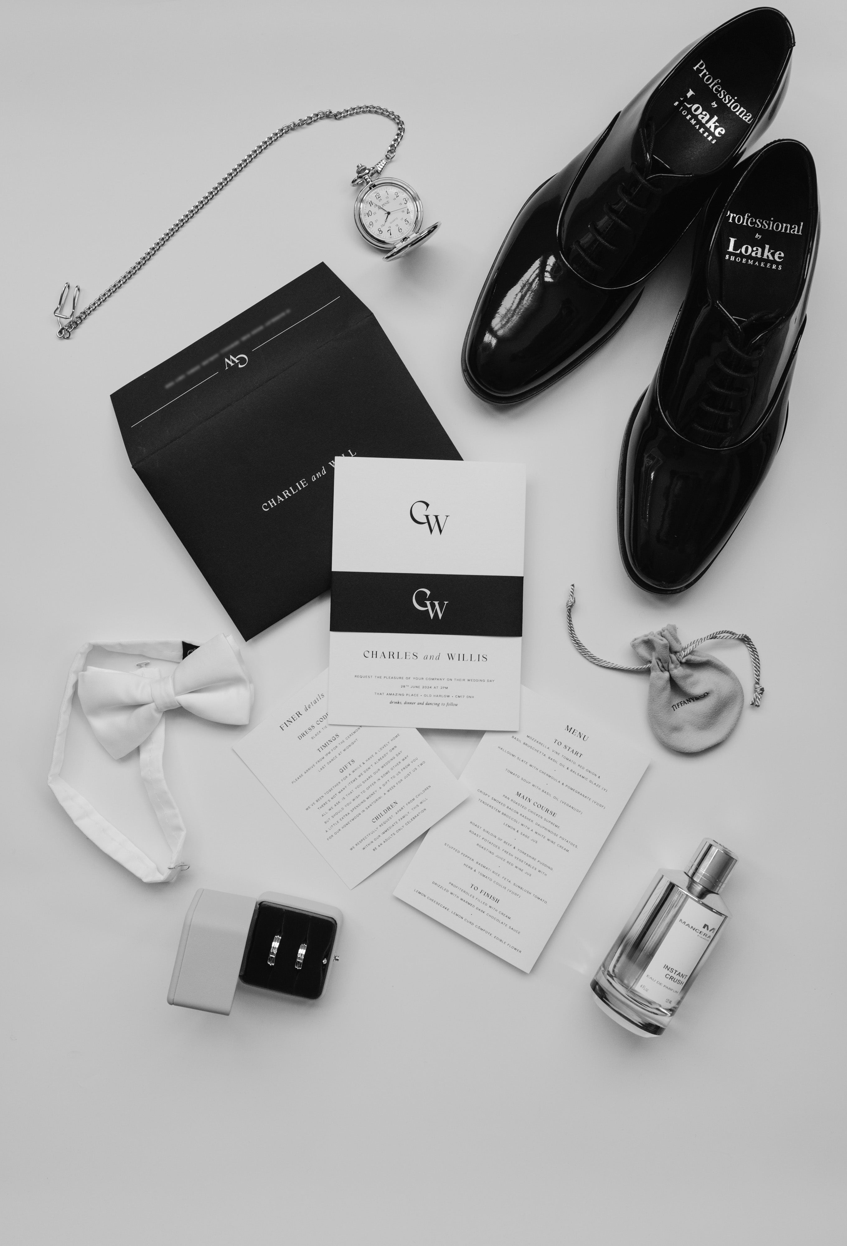 Black and white wedding invite suite, pcitured as a falt lay with black belly band, grooms shoes, aftershave and wedding rings