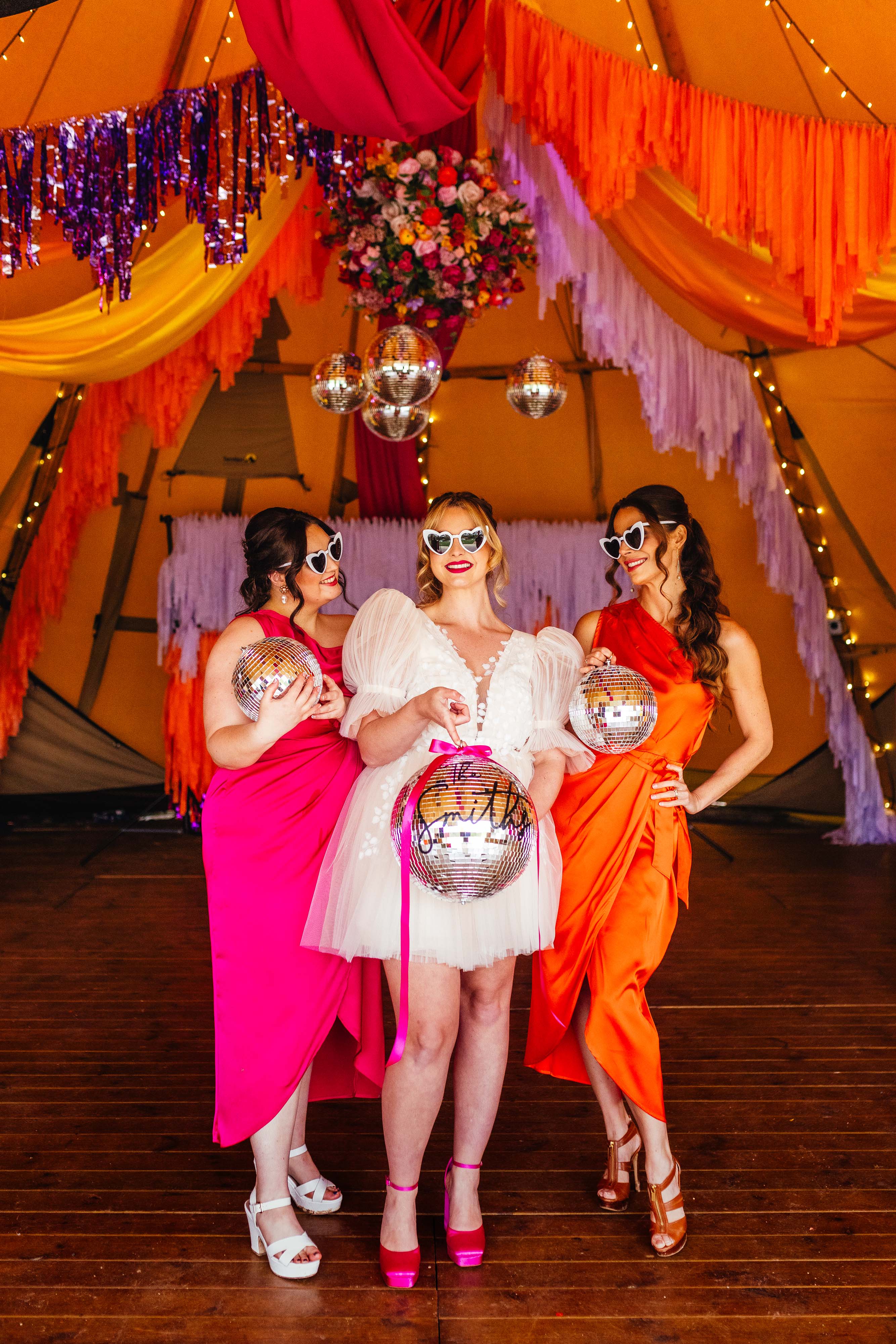 Disco style wedding styling with bright oranges, and pinks, bride holding a disco ball, with bridesmaid either side in neon pink and neon orange dresses, with streamers hanging from the ceiling