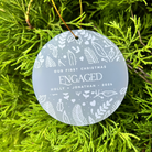 A round frosted glass Christmas ornament with white etched botanical patterns and the text 'Our First Christmas Engaged Holly + Jonathan 2026', hanging from a gold string amidst bright green pine needles.