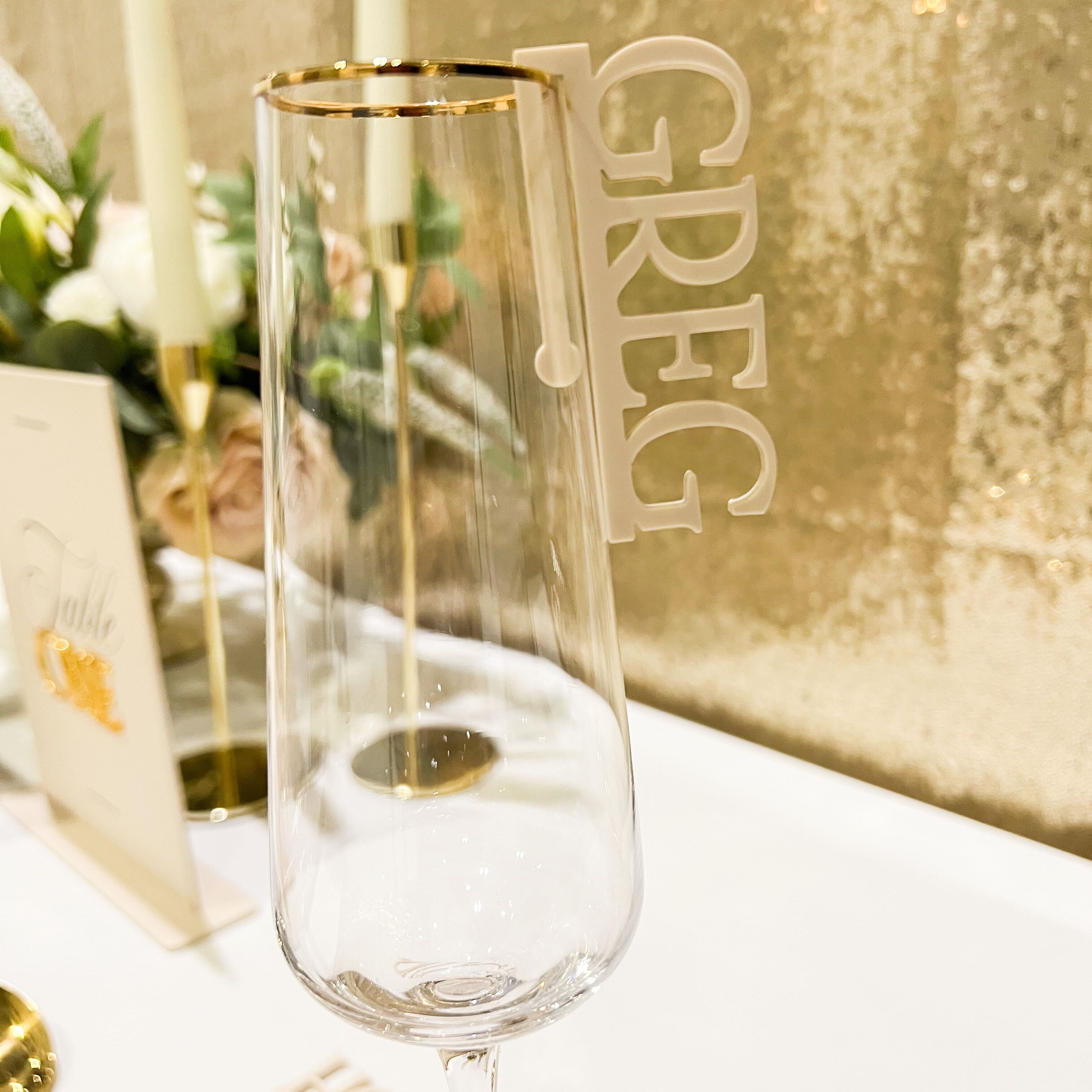 ACRYLIC WINE GLASS CHARM CLIPPED TO THE SIDE OF A CHAMPAGNE FLUTE SPELLING THE NAME GREG