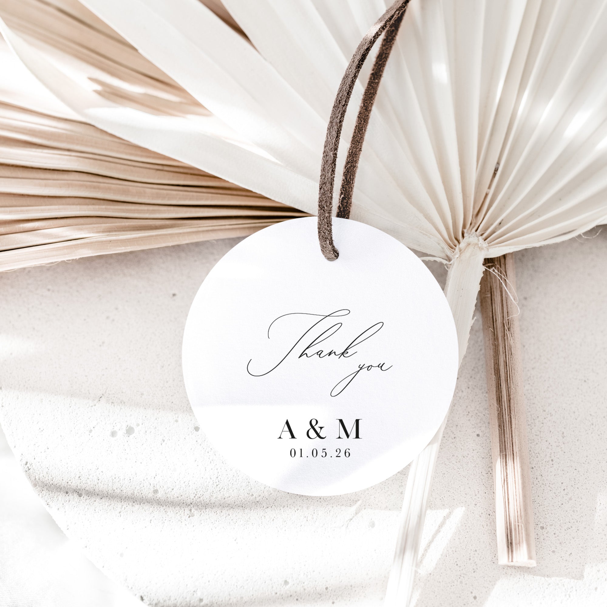 Round white tag with 'Thank you A&M 01.05.26' text, tied to dried palm leaves on a textured surface.