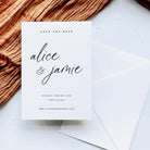 A white save the date card shown above a white diamond flap envelope on a white background, next to copper coloured fabric