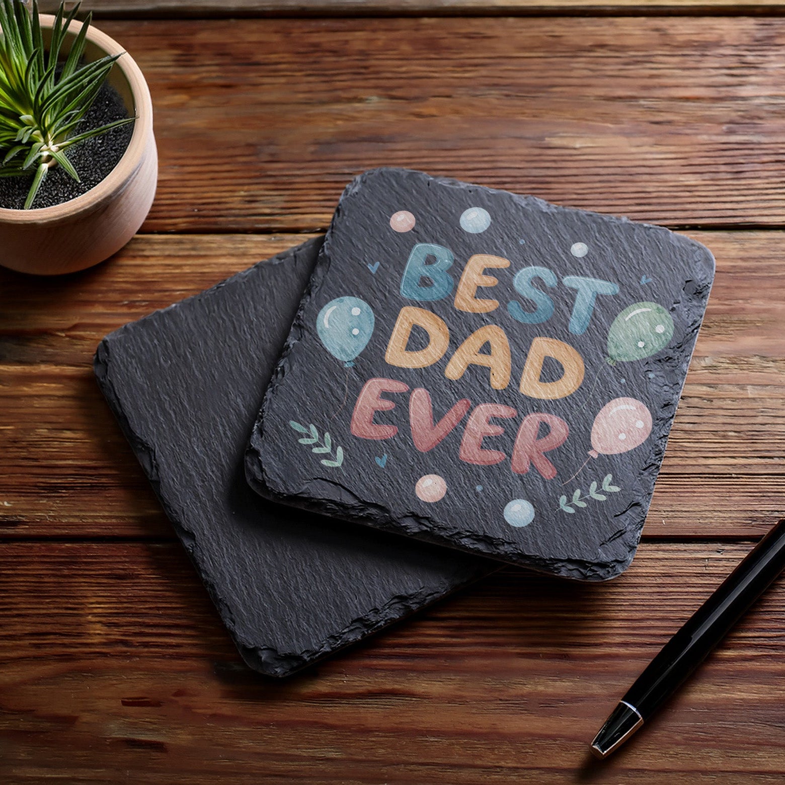 Slate coaster with 'Best Dad Ever' text on a wooden surface with a pen and plant.