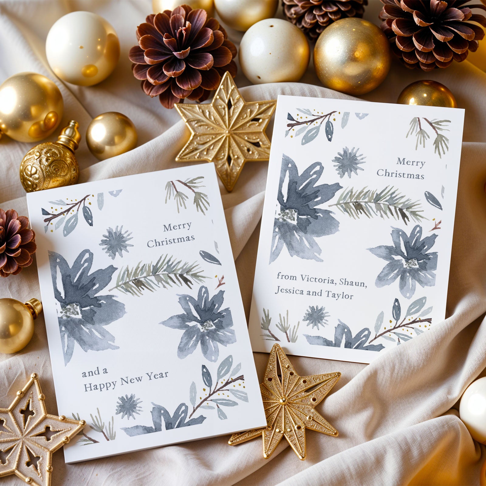 Two Christmas cards with floral designs surrounded by gold ornaments and pine cones on a beige fabric background.