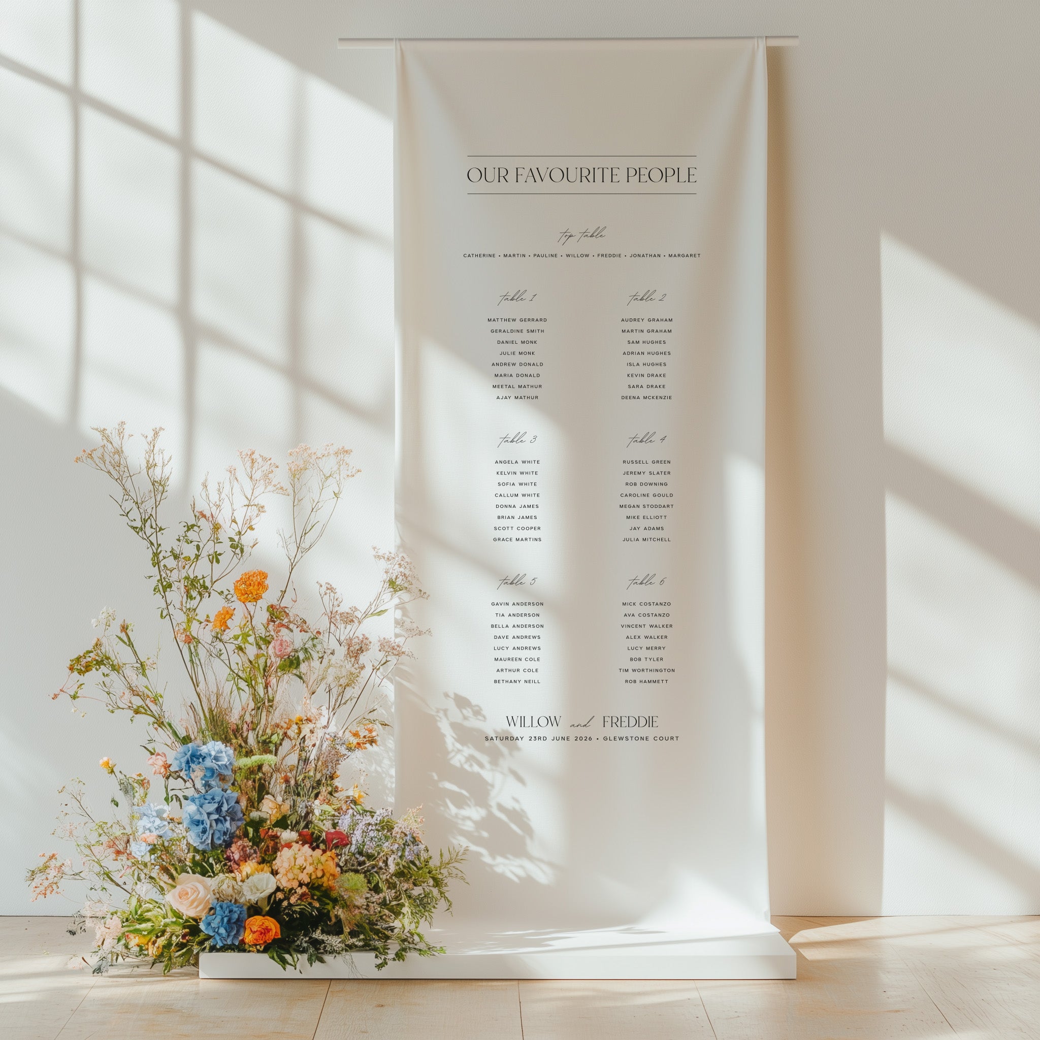 Floral arrangement in a vase next to a seating chart on a white wall with sunlight filtering through.