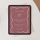 A tablet displaying a custom 'Save the Date' wedding invitation card with a dusty rose background and white script text, styled on top of a white envelope and a beige textured surface.