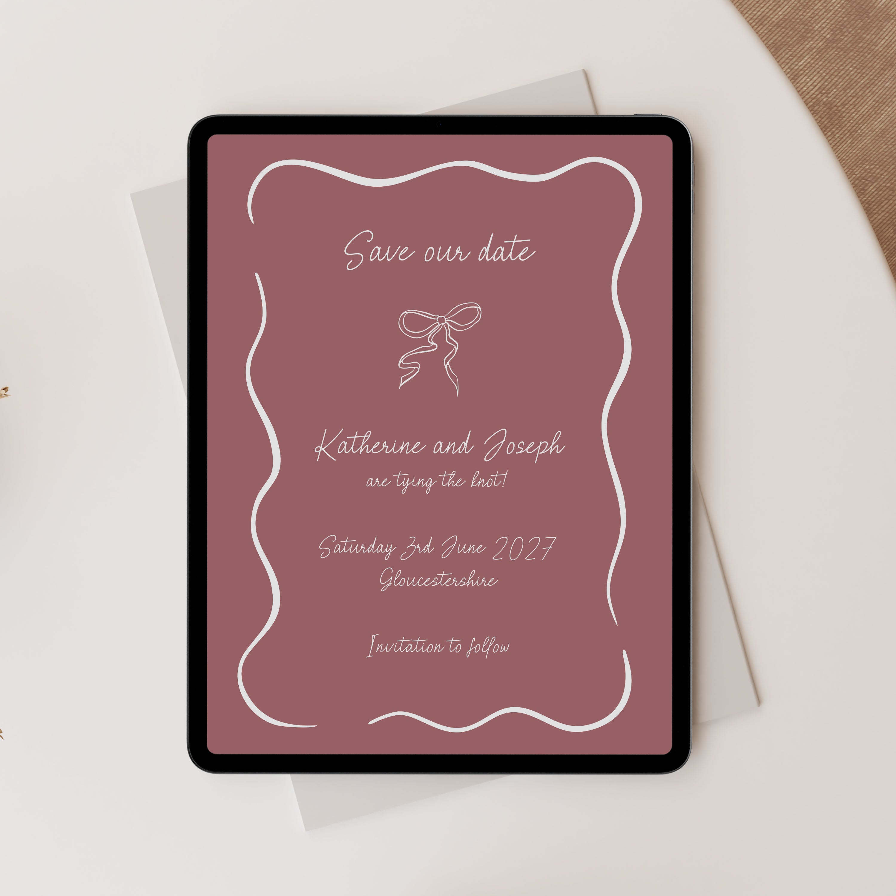 A tablet displaying a custom 'Save the Date' wedding invitation card with a dusty rose background and white script text, styled on top of a white envelope and a beige textured surface.