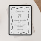 A digital 'Save our date' with elegant black typography and a bow illustration on a white background, styled on top of a light envelope over a cream surface with a beige fabric corner visible.