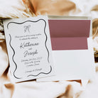 A wedding invitation card with elegant black script and border details, displayed next to a matching white envelope with a dusty rose card insert, set against a soft beige fabric background.