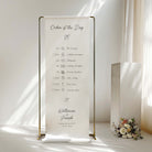 A vertical white fabric wedding order-of-the-day sign with gold metal frame stands on a white floor in a softly lit room. The sign displays a printed timeline from the wedding ceremony to dinner, with the names Katherine and Joseph and the date printed at the bottom.