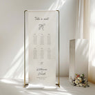 A freestanding vertical wedding seating chart sign in fabric on a gold metal frame, displayed on a table in a softly lit room with sunlight and a small floral arrangement nearby.