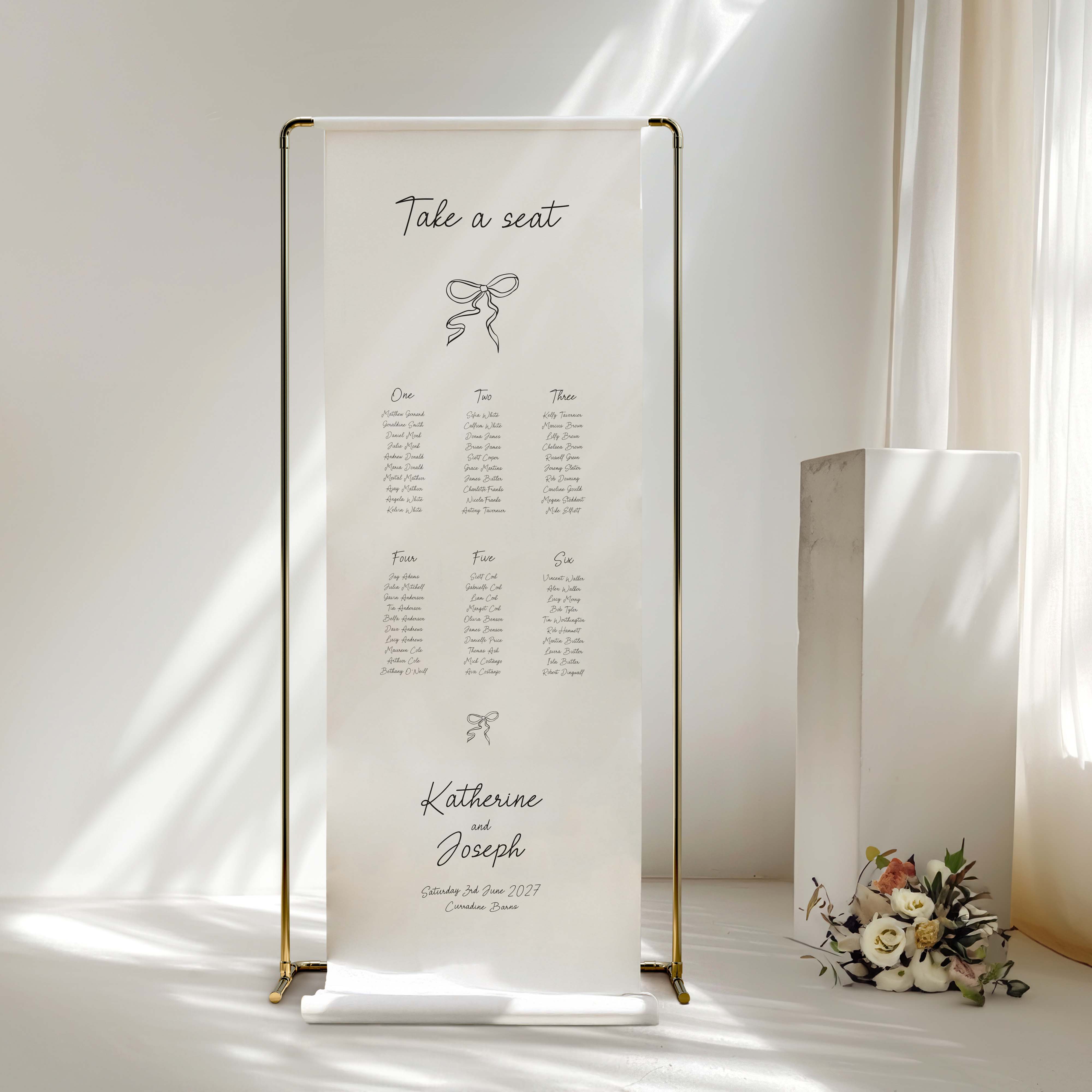 A freestanding vertical wedding seating chart sign in fabric on a gold metal frame, displayed on a table in a softly lit room with sunlight and a small floral arrangement nearby.