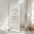 A vertical white fabric wedding welcome sign on a slim gold metal stand, displayed in a bright, sunlit room next to a white pedestal and a small floral arrangement.