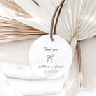 A round white wedding thank-you tag with a brown string, printed with 'Thank you', a small bow illustration, and the names Katherine + Joseph plus the date 03.06.