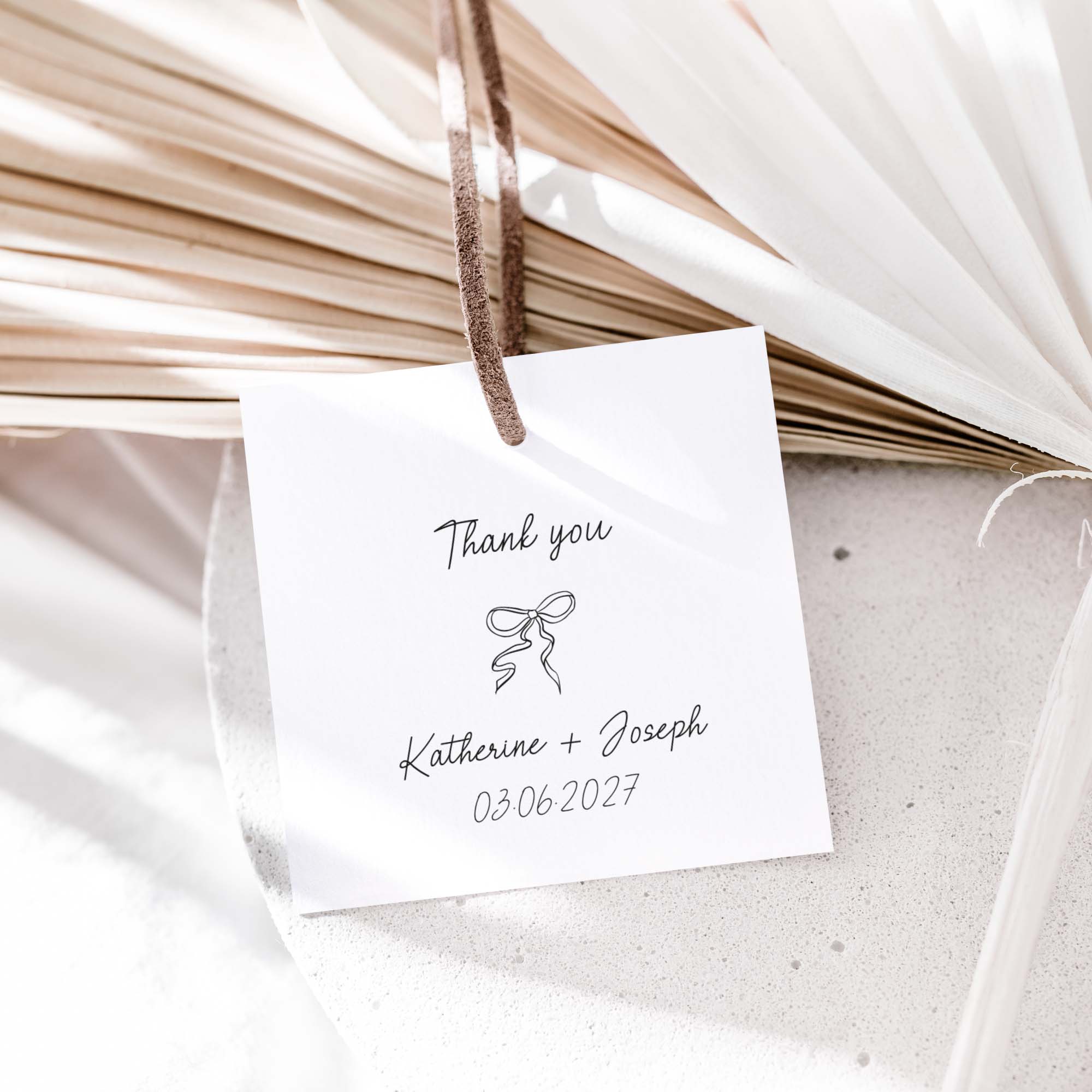 A small white wedding favor tag with black script reading 'Thank you' and 'Katherine + Joseph 03.06.