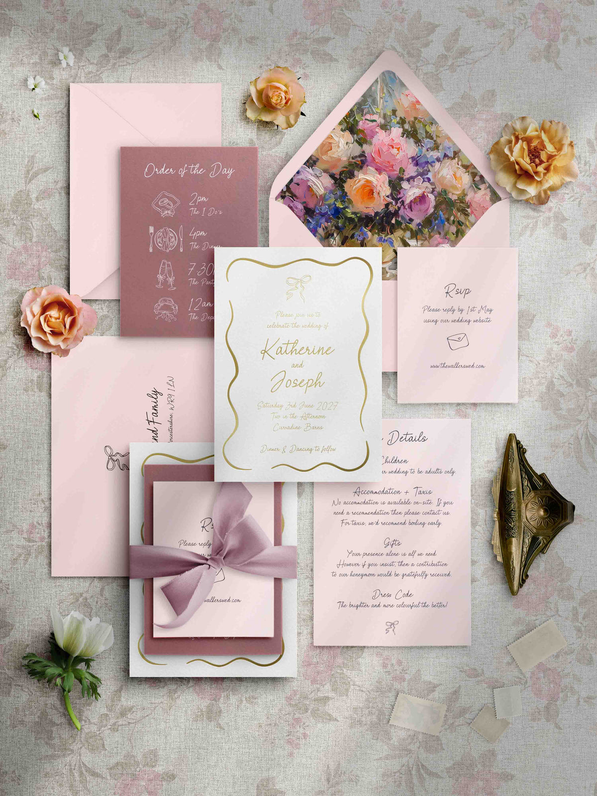 A coordinated wedding stationery flat lay on a textured light gray surface, including a floral invitation card, pink envelopes, a details card, a rose invitation, and a gift card tied with a mauve ribbon. Decorative flowers and a gold pocket watch surround the stationery.