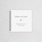 A personalized spiral-bound wedding guest book with a white cover, black wire binding, and custom printed text reading 'Katherine and Joseph' with a small bow illustration, displayed on a light gray textured fabric background.