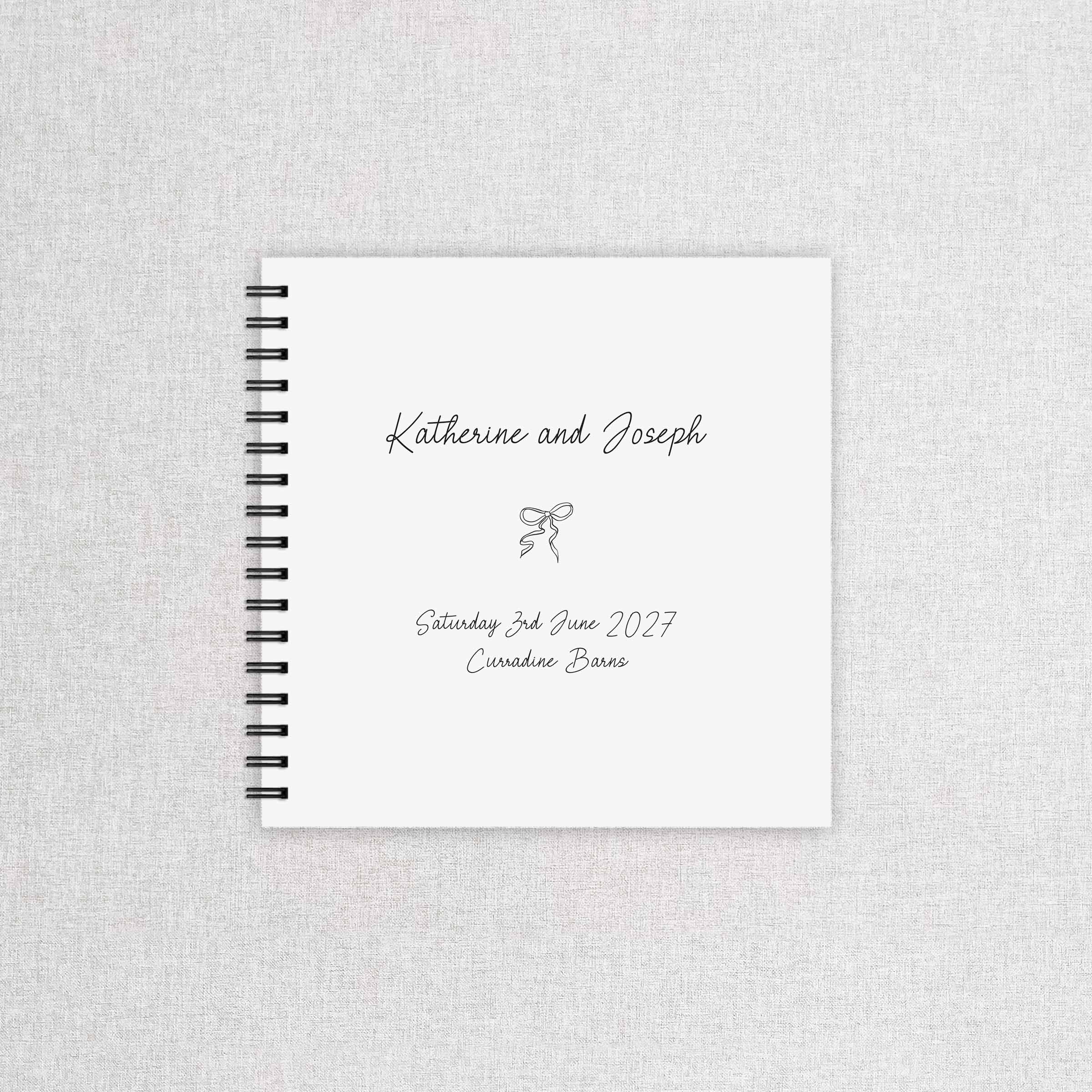 A personalized spiral-bound wedding guest book with a white cover, black wire binding, and custom printed text reading 'Katherine and Joseph' with a small bow illustration, displayed on a light gray textured fabric background.