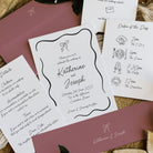 A styled flat-lay of wedding stationery including a white invitation card with black script text, an order of the day card, and matching dusty rose envelopes on a neutral surface.