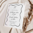 A white wedding order of service booklet with elegant black script text and a decorative wavy border, styled on draped cream fabric with dried pampas grass.