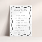 A vertical white wedding order of the day board with a black decorative border and handwritten-style event timeline, shown against a light beige wall and surface.
