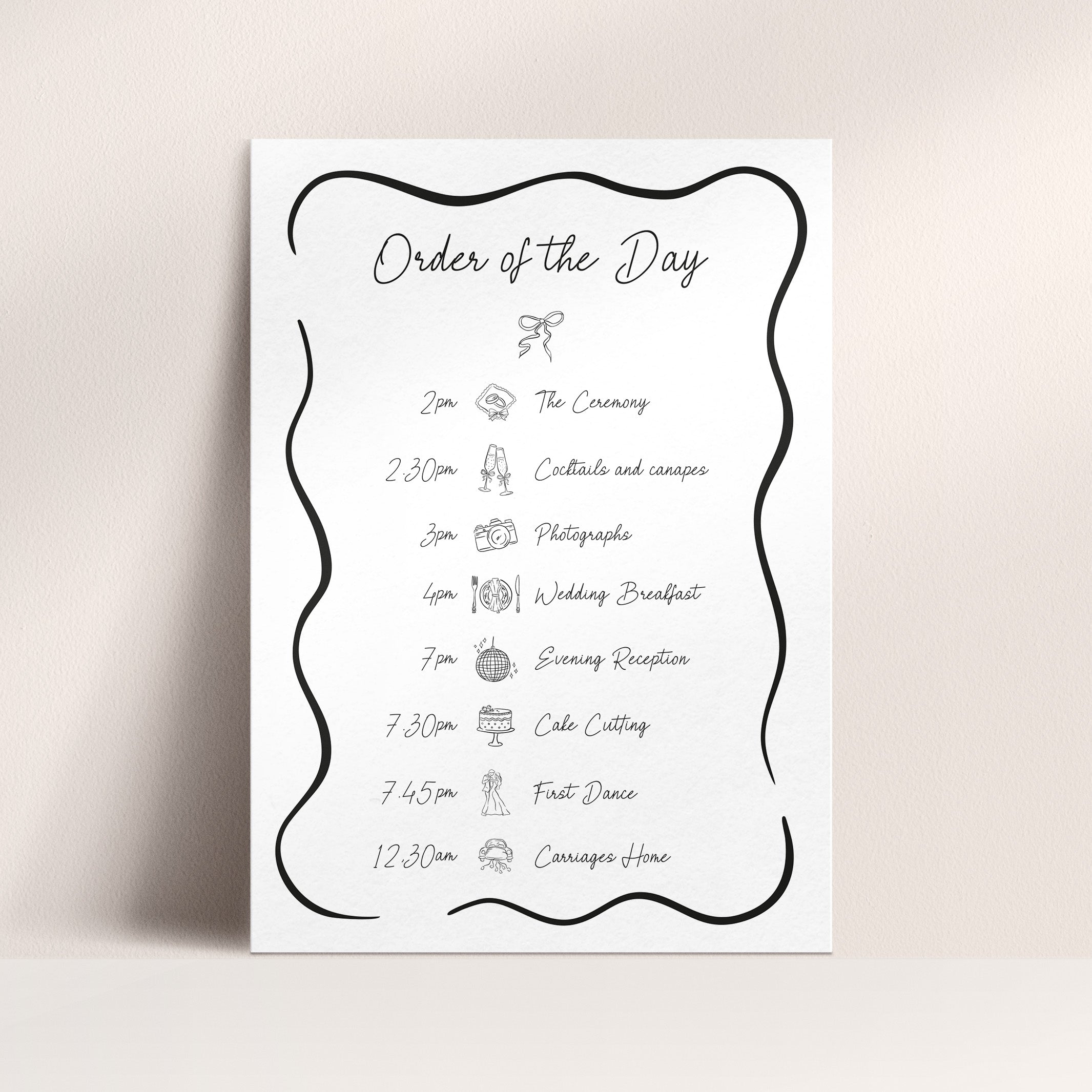 A vertical white wedding order of the day board with a black decorative border and handwritten-style event timeline, shown against a light beige wall and surface.