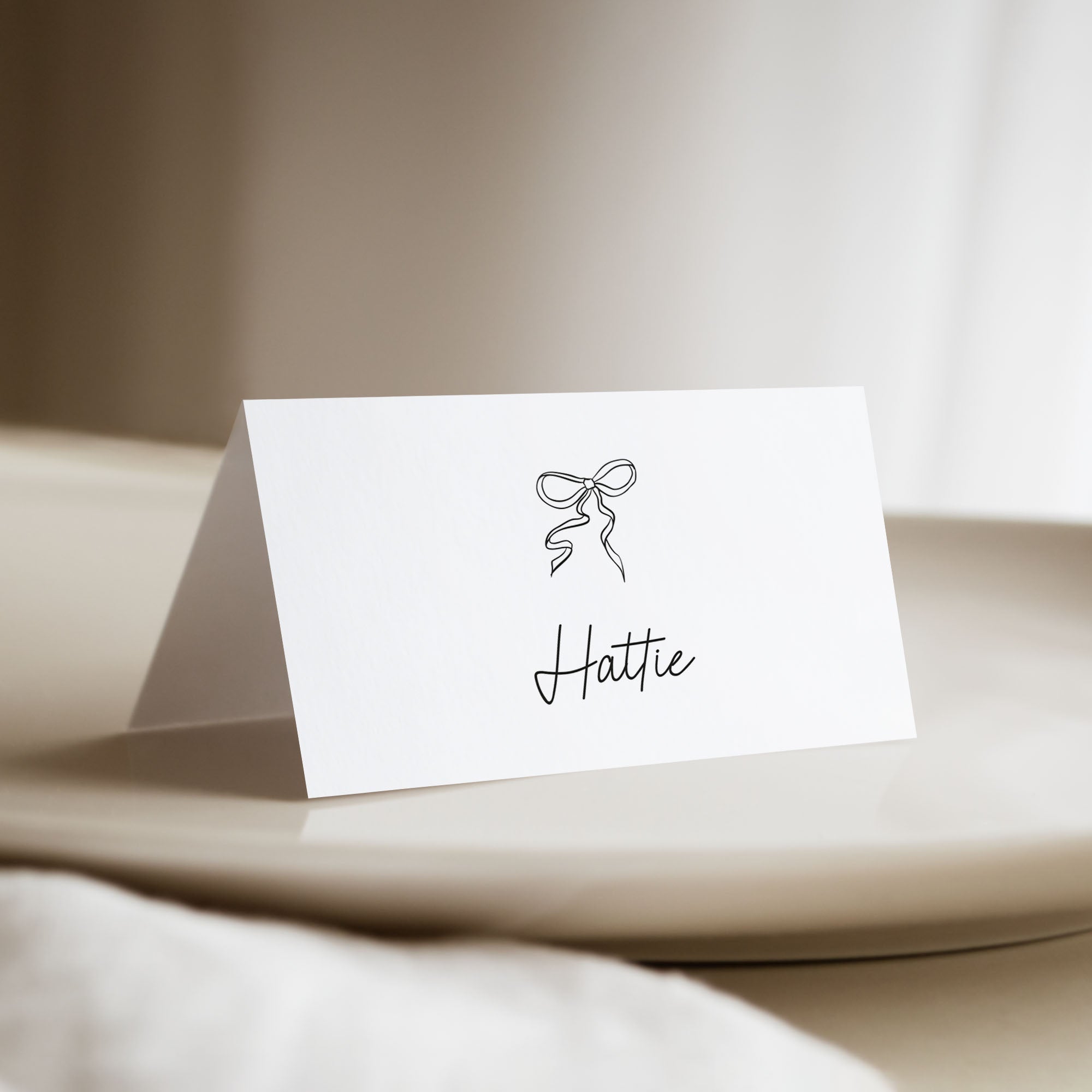 A white standing place card with a black ribbon icon and the name 'Hattie' in script, displayed on a beige table in a softly lit indoor setting.
