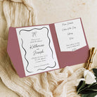 A wedding invitation suite displayed in an open pocket folder with a dusty rose cover, styled on cream fabric with a gold pen and a white rose.