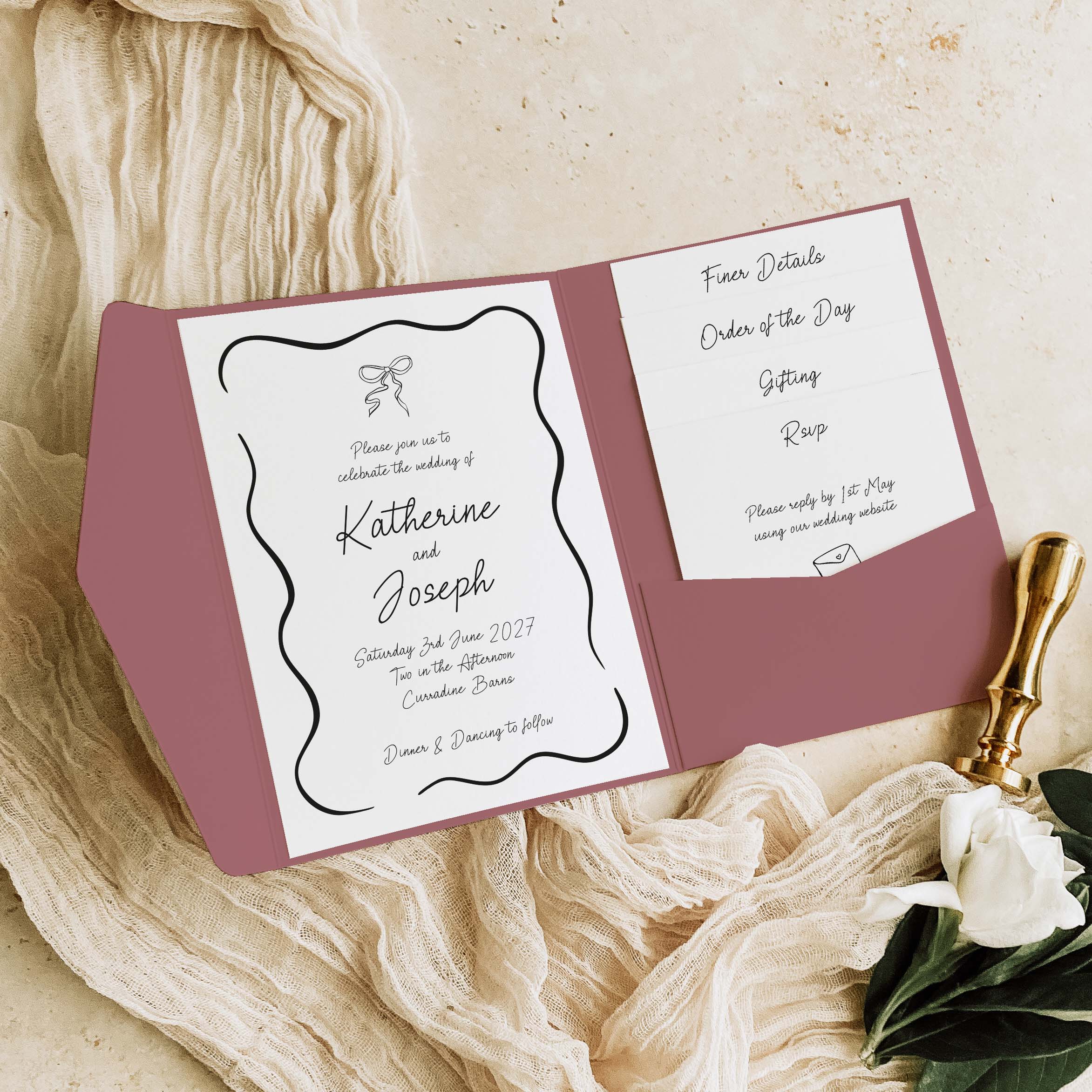 A wedding invitation suite displayed in an open pocket folder with a dusty rose cover, styled on cream fabric with a gold pen and a white rose.