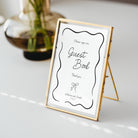 A gold-framed standing guest book sign with a white insert and black script text, photographed on a light tabletop with a blurred glass vase in the background.