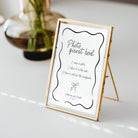 A gold-framed photo guest book sign on a white A-frame stand, placed on a light surface with a blurred glass vase in the background.