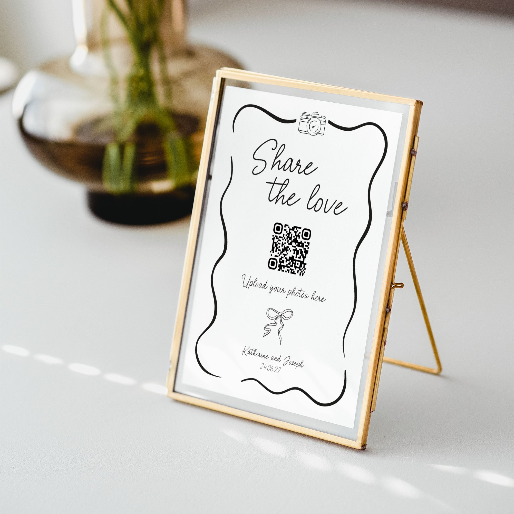 A gold-framed vertical photo frame stand holding a white sign that reads 'Share the love' with a QR code placeholder, photographed on a light tabletop with a blurred glass vase in the background.