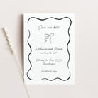 A white 'Save our date' card with a black wavy border and elegant script text, photographed on a light beige background with a dried wheat stem to the left.